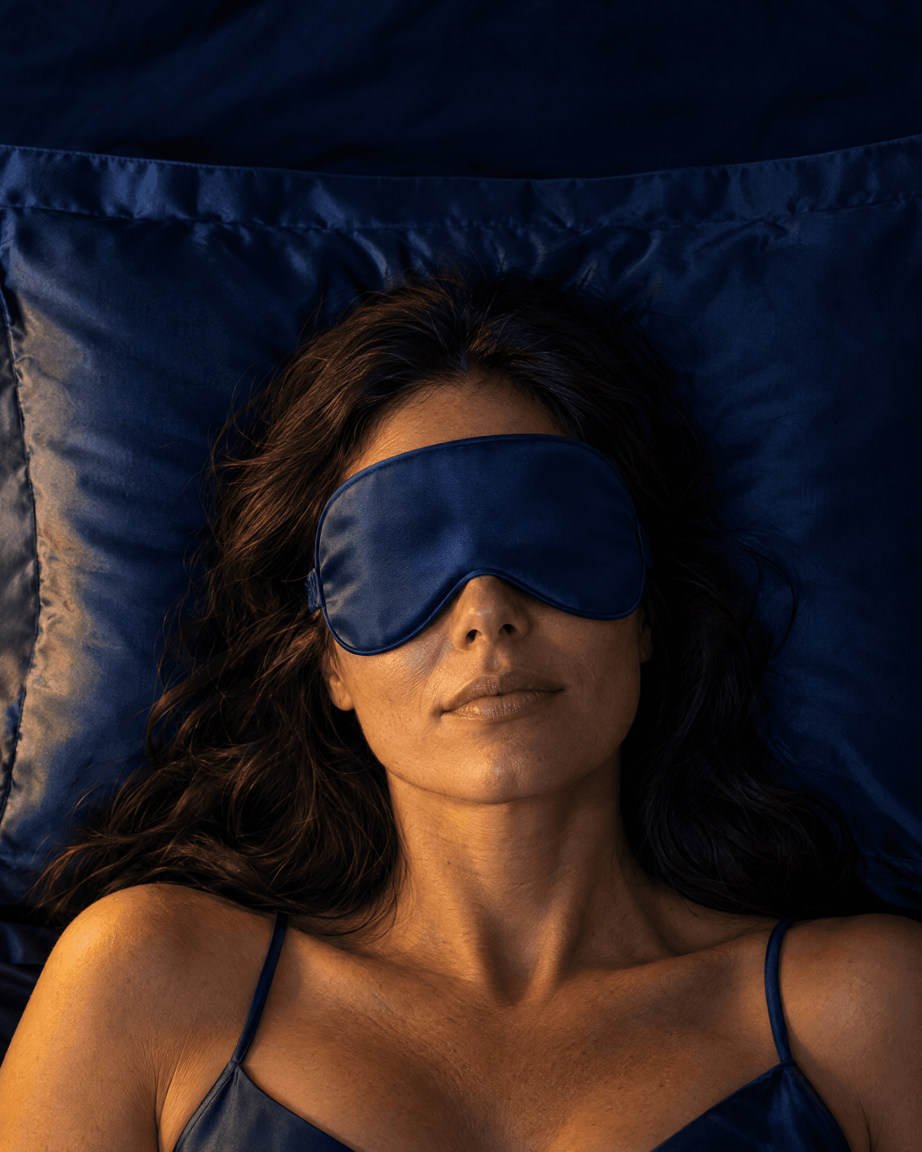 A woman lying on a blue silk pillowcase wearing a contoured silk eye mask